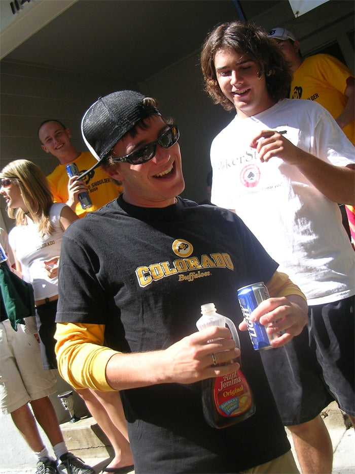 Alex Kanzler wearing sunglasses and a black shirt with yellow text, holding a bottle and can, with others in the background.