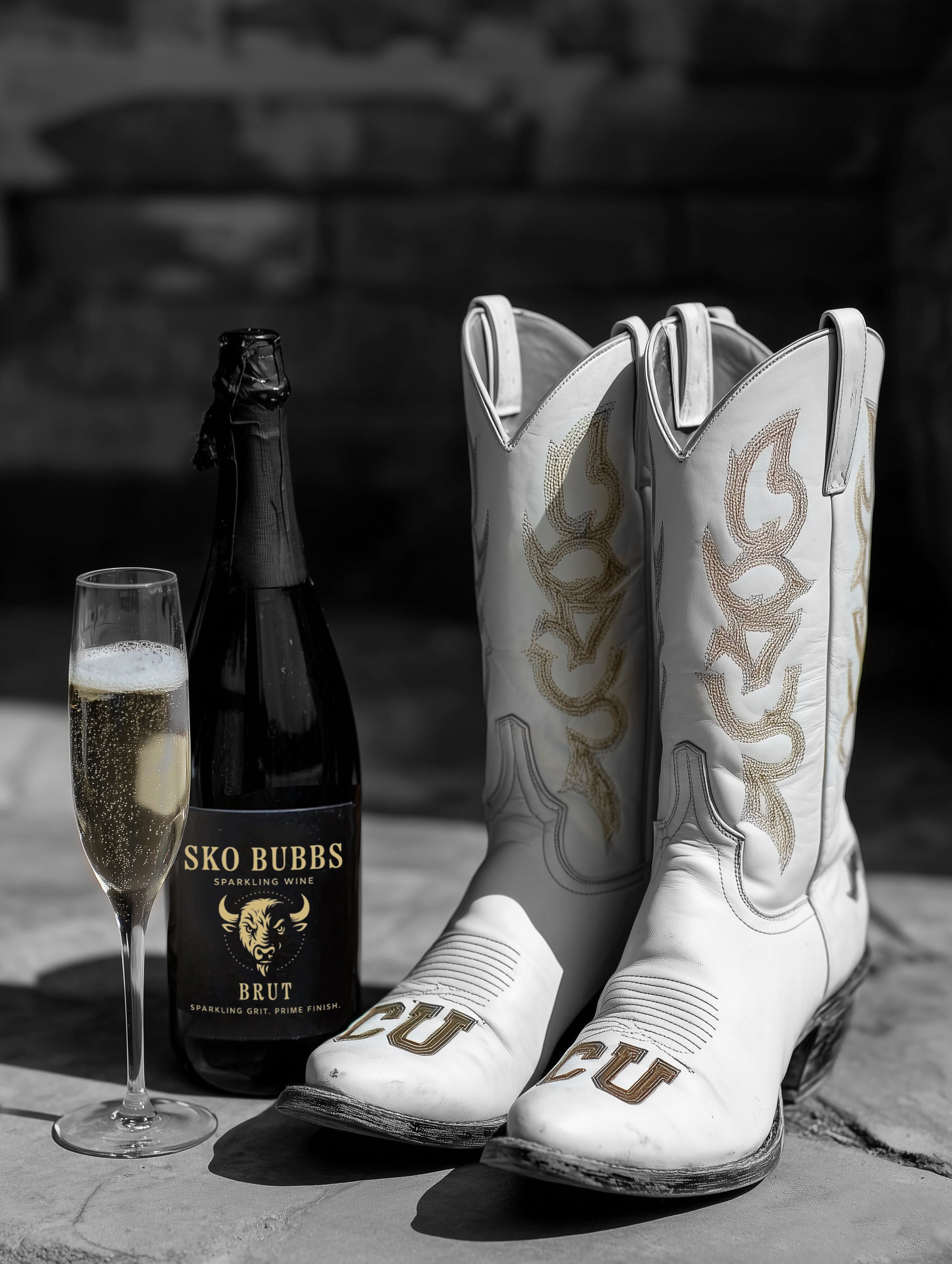 White cowboy boots with flame design, bottle of Sko Bubbs sparking wine, and glass of champagne on a stone surface.